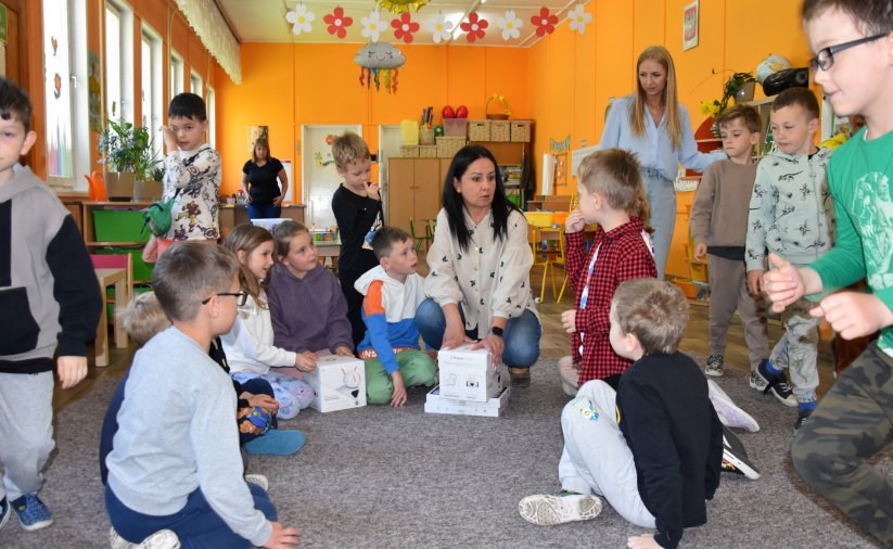 Grupa przedszkolaków w dużej sali, siedzących na podłodze i rozpakowujących z pudełek roboty i tablety