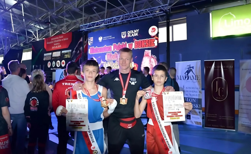 Dwóch młodych zawodników w sportowych strojach, z dyplomami i medalami, pomiędzy nimi trener