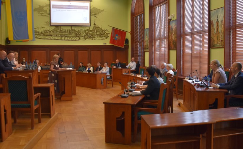Duża sala z parkietem i drewnianymi stołami, przy stołąch siedzą radni