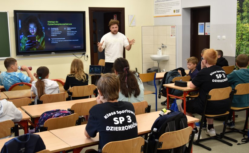 Uczniowie siedzący w ławkach szkolnych, przy interaktywnej tablicy dorosła osoba w trakcie prezentacji