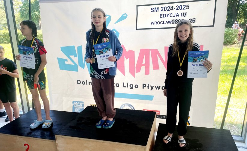 Laila Lachowicz na najwyższym stopniu podium z medalem i dyplomem, po bokach dwie pływaczki