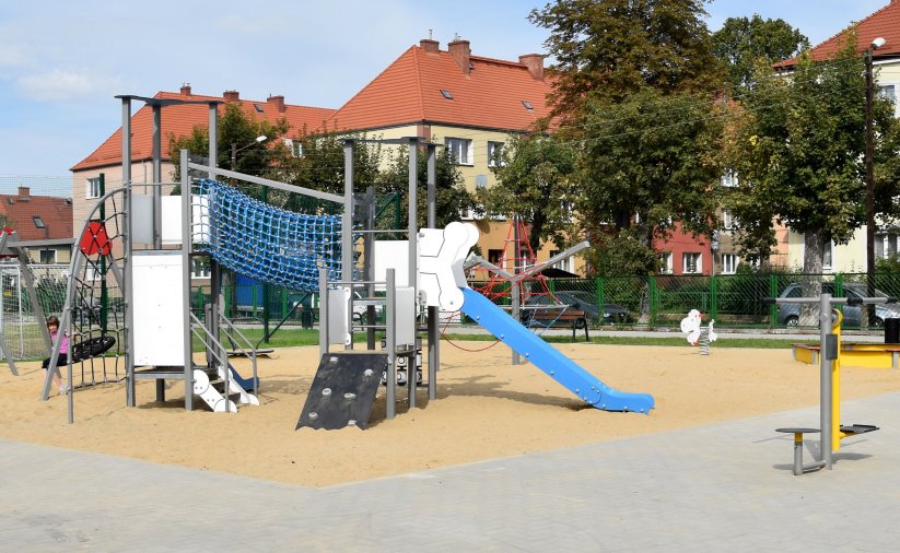 Plac z urządzeniami do zabawy zamontowanymi na piaszczystym podłożu, w tle budynki wielorodzinne