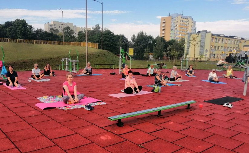 Kilkanaście osób siedzących na matach do ćwiczeń na terenie zewnętrzej siłowni, za nimi zieleń i bloki osiedlowe