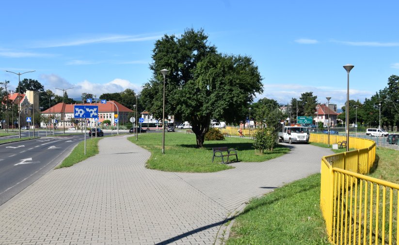 Droga w mieście, obok chodnik i teren zielony z dużym drzewem, po prawej żółte barierki