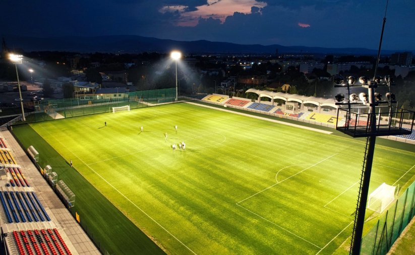 Stadion piłkarski nocą, oświetlony, z trybunami po bokach, na muraiwe grupka piłkarzy