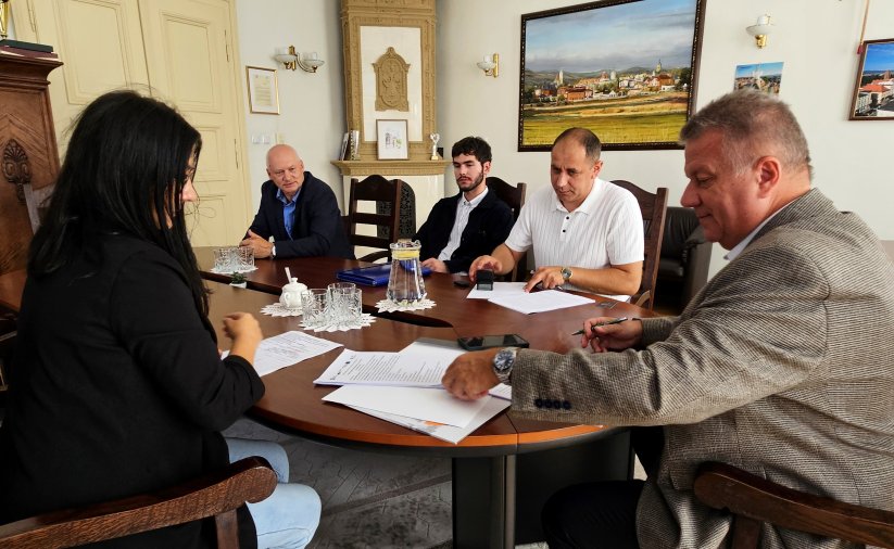 Zdjęcie przedstawiające siedzących przy stole czterech mężczyzn i kobietę. Dwóch z mężczyzn podpisuje dokumenty