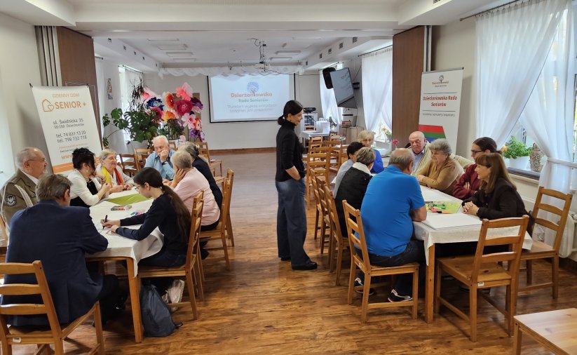 Dwie grupy ludzi przy stołach w sali multimedialnej w Centrum Seniora, pośrodku prowadząca warsztaty pani psycholog