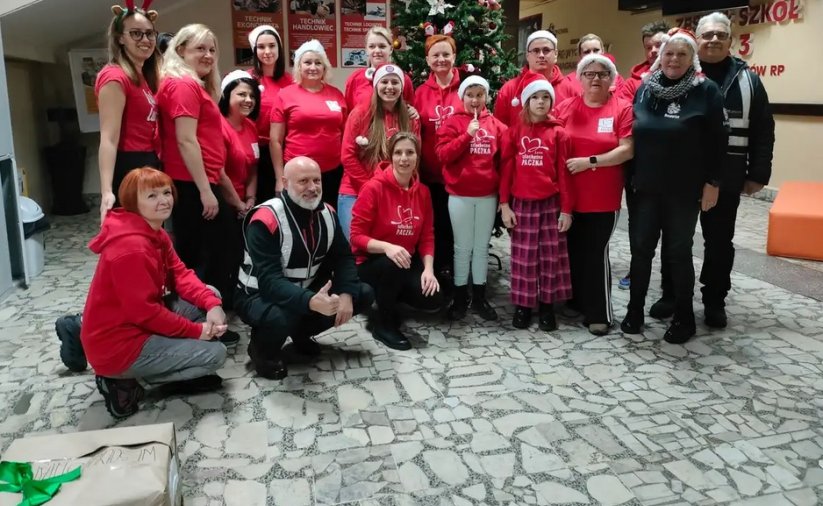 Grupa wolontariuszy Szlechetniej paczki w czerwonych koszulkach i czapkach św. Mikołaja stoi w korytarzu Zespołu Szkół nr 3 w Dzierżoniowie