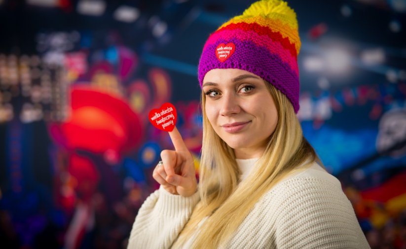 Blondynka w kolorowej czapce s zerduszkiem WOŚP trzyma na palcu serduszko WOŚP