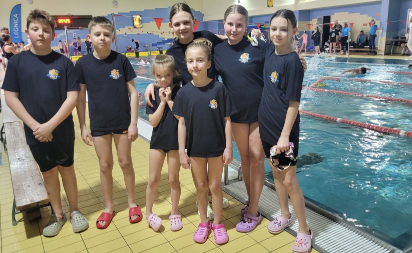 Siedmioro młodych pływaków w jednakowych granatowych koszulkach przy krawędzi basenu