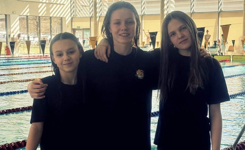 Trzy młode zawodniczki w czarnych koszulkach na tle basenu