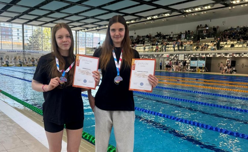 Dwie dziewczyny z medalami i dyplomami stoją przy krawędzi basenu