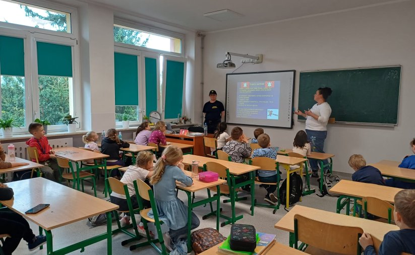 Sala w szkole, dzieci siedzą w ławkach, patrzą w kierunku tablicy, przy niej dwie kobiety, jedna z nich wskazuje ręką na tablicę