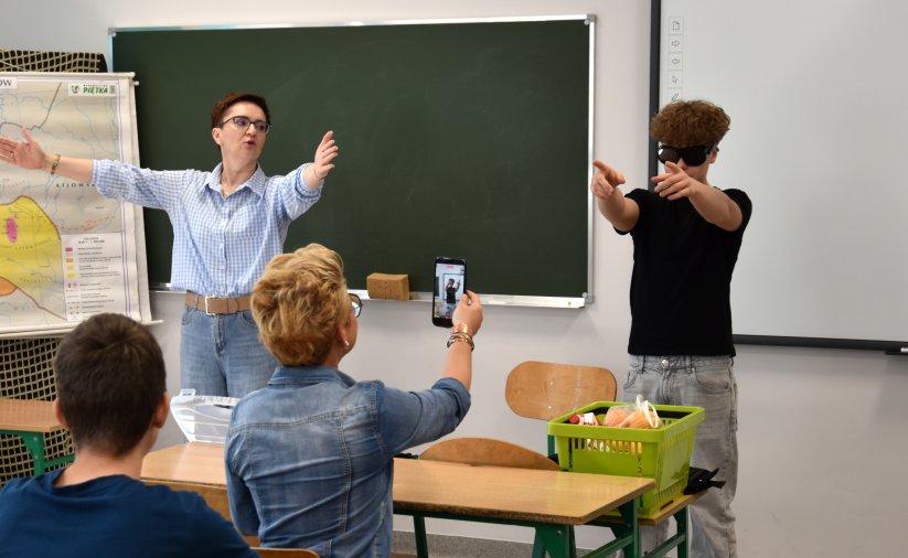Prowadząca zajęcia przy szkolnej tablicy z rozłożonymi rękoma, obok uczeń w ciemnych okularach wskazujący na drugi koniec sali