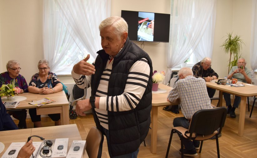 Senior z opaską na ręce, za nim przy stołach siedzi grupa seniorek i seniorów