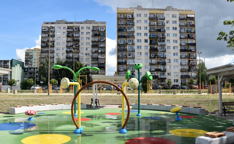 Wodny plac zabaw z zielona nawierzchnią i z urządzeniami tryskającymi wodę, w tle dwa wieżowce