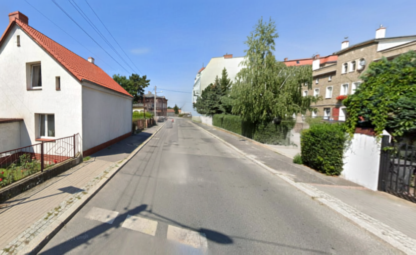 Droga asfaltowa, po lewej biały domek jednorodzinny, po praweej drzewa i domy jednorodzinne