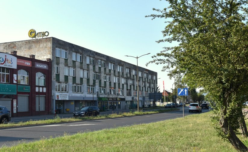 Długi trzypiętrowy budynek położony przy drodze, po prawej stronie drzewo