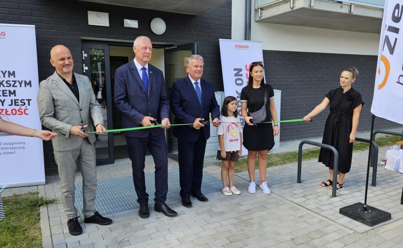 Trzech mężczyzn w garniturach i mama z dzieckiem przecinają zielona wstęgę przed wejściem do budynku