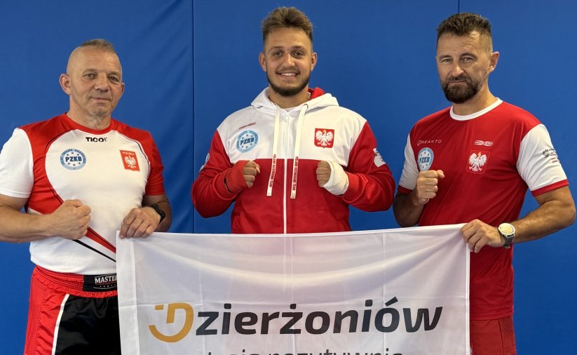 Maksymilian Pelej w biało-czerwonym dresie. Po bokach trenerzy trzymający flagę z logotypem Dzierżoniowa