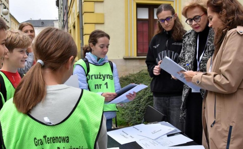 Uczniowie w zielonych kamizelkach z napisem "Gra terenowa" przy stolu ustawionym na ulicy, obok trójka organizatorów konkursu