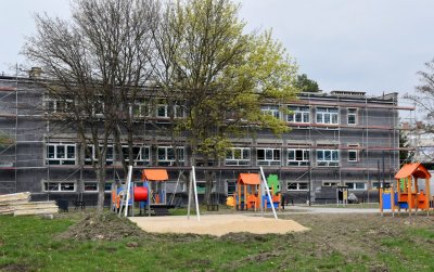 Plac zabaw z urządzeniami i piaskową nawierzchnią, w tle budynek w trakcie remontu