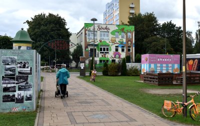 Kobieta prwadząca wózek, idąca aleją prowadzącą do żłobka, na ścianie budynku kolrowy mural z sowami