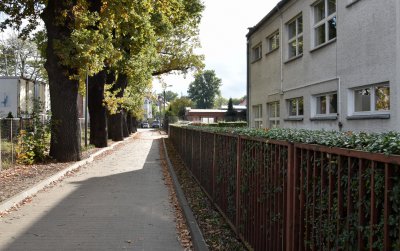 Droga z kostki betonowej, po prawej stronie ogrodzenie, żywopłot i fragment budynku