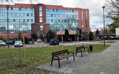 Dwie ławki rzy chodniku, w drugim planie czerwony, mocno przeszklony budynek