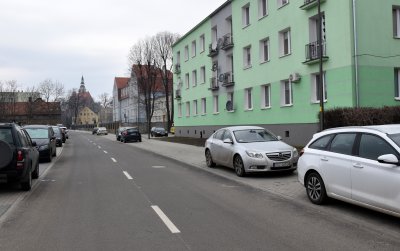 Nowa droga asfaltowa, na chodnikach zaparkowane samochody, po prawej budynek wielorodzinny