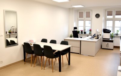 Duże jasne pomieszczenie, stół ustawiony przy ścianie, przy oknach biurko