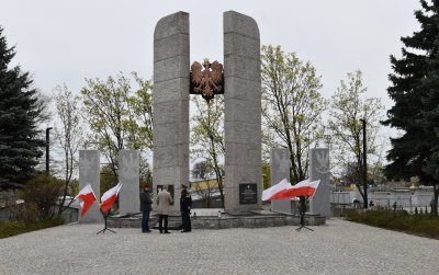 Pomnik, przed nim flagi, po bokach drzewa iglaste