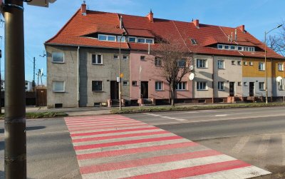 Przejście dla pieszych w białe i czerwone pasy, za nim długa kamienica