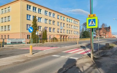 Szeroka droga, białoczerwone przejścia dla pieszych i duży czteropiętrowy żółty budynek