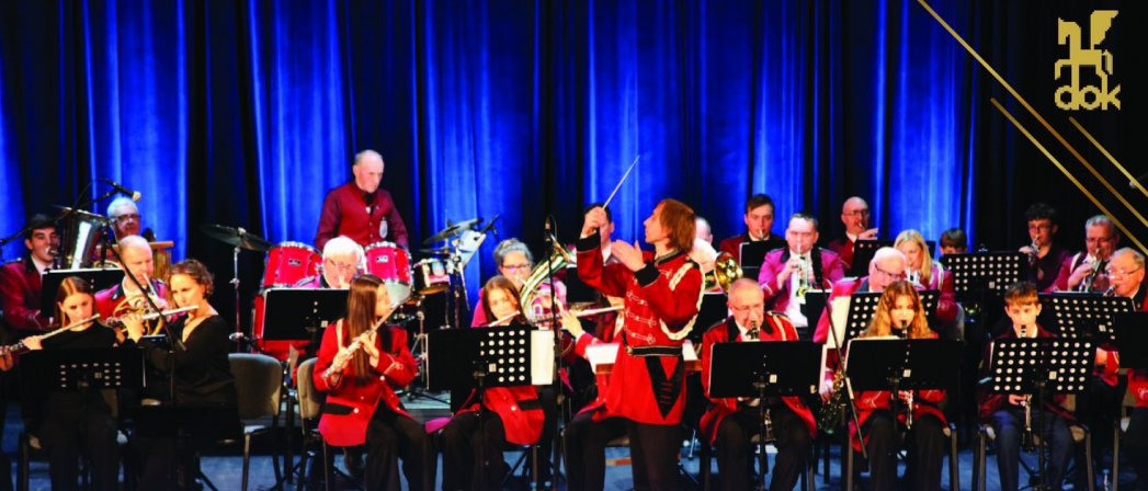 Orkiestra dęta na scenie, czerwone stroje muzyków, niebieskie tło
