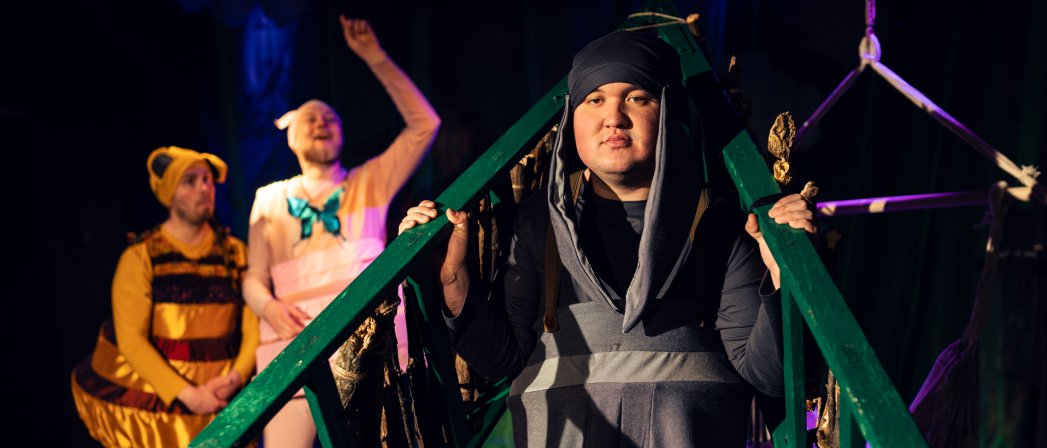 Trójka aktorów na scenie, jeden przebrany za pszczołę, ciemne tło