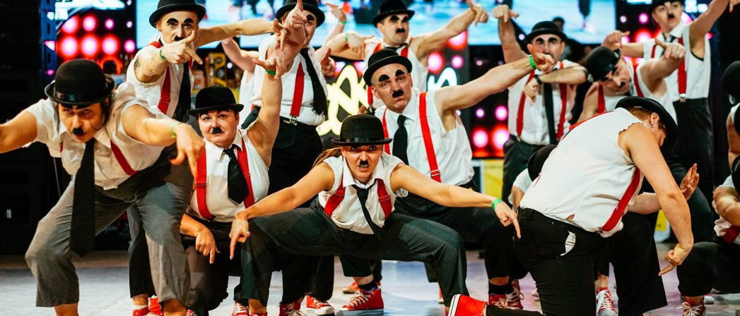 Grupa tancerzy, kobiety i mężczyźni na scenie w róznych pozach, ubrani i ucharakteryzowani jak Charlie Chaplin, brytyjski aktor