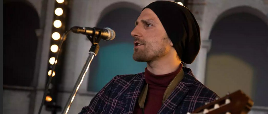 Mężczyzna w czarnej czapce, z gitarą w rękach śpiewający do mikrofonu, Fot. https://polskanews.pl