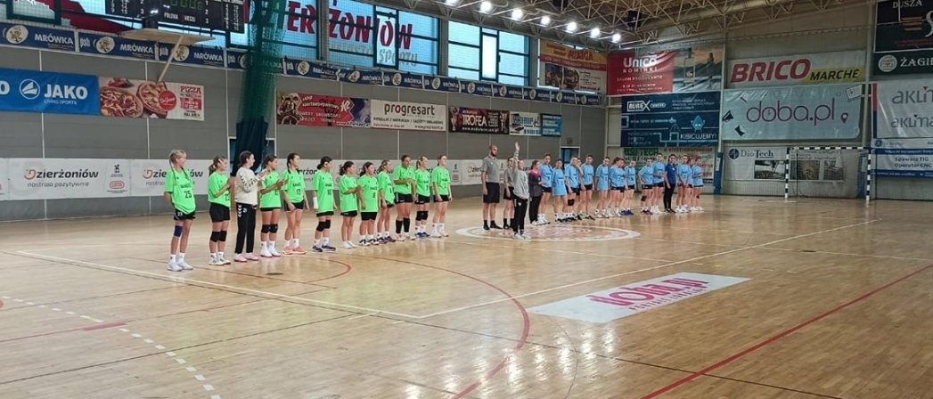 Dwie drużyny dziewczęce stojące równo na środku drewnianego parkietu w hali sportowej