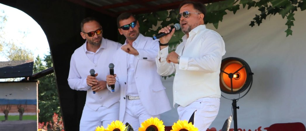 Trzech mężaczyzn w białych strojach śpiewa na niewielkiej scenie
