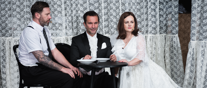 Kobieta w białej sukni i dwóch meżczyzn siędzą przy małym stoliku, z tyłu wiszące firanki