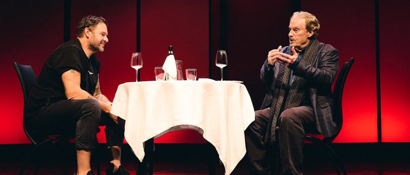 Spektakl, dwóch aktorów rozmawia siedząc przy stoliku z białym obrusem 