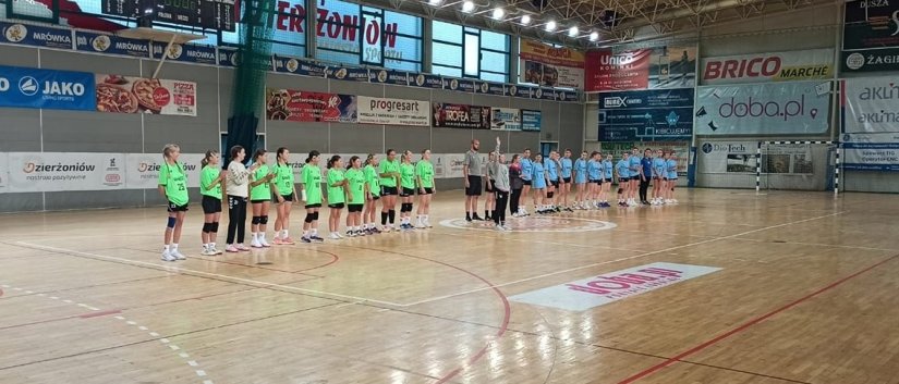 Dwie drużyny dziewczęce stoją równo na środku drewnianego parkietu w hali sportowej